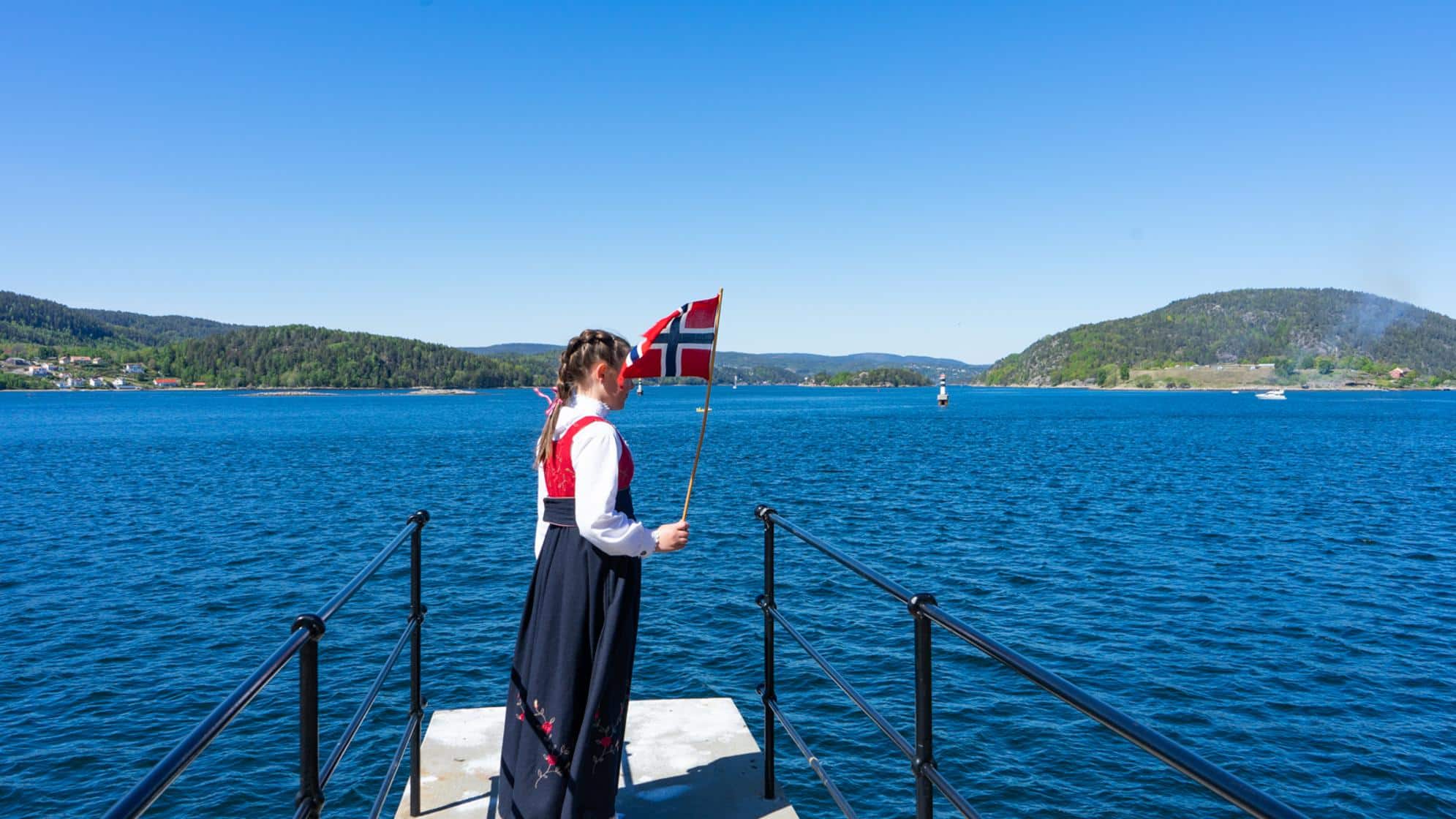 Dame som står i bunad og ser utover fjorden med flagg på 17.mai