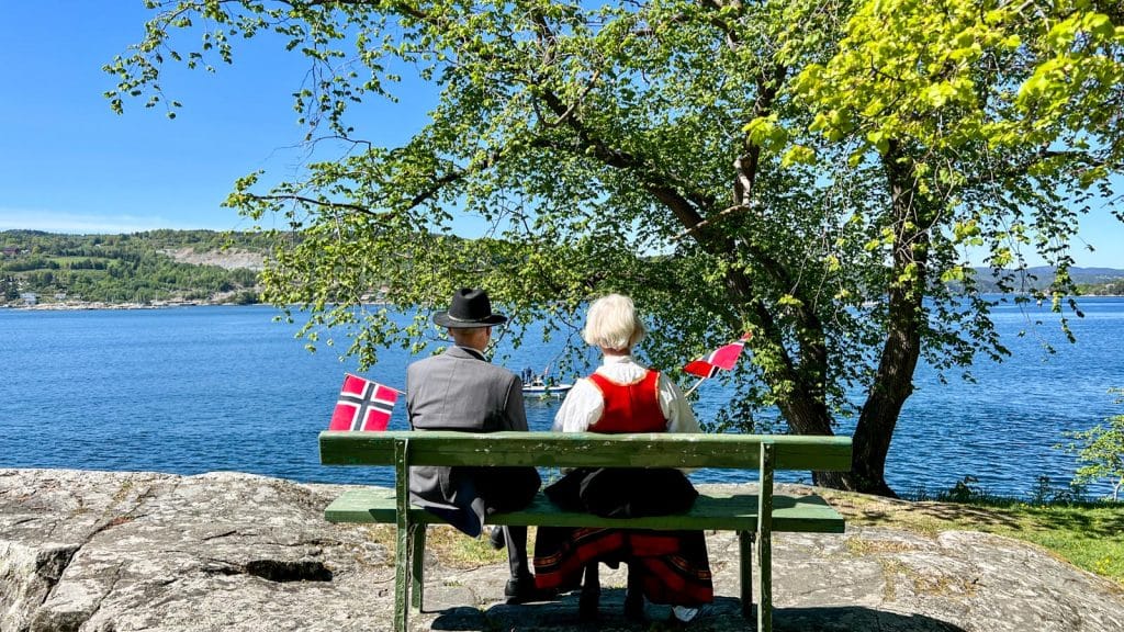 Et par i bunad som sitter på en benk på 17.mai med flagg