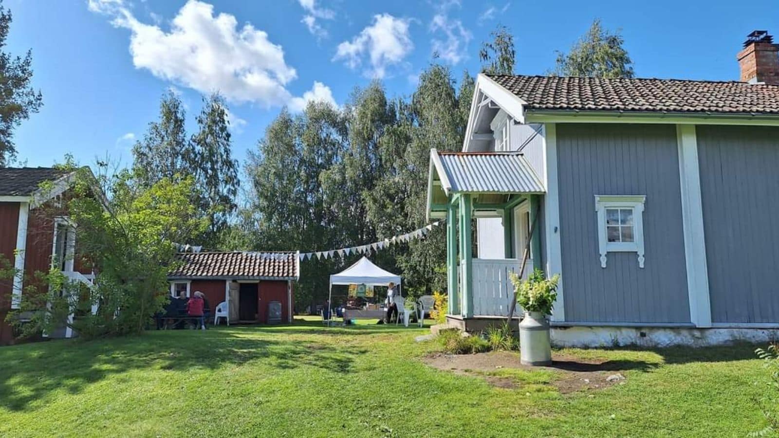 lyseblått gammelt hus med et stabbur og et lite rødt hys med mennesker foran