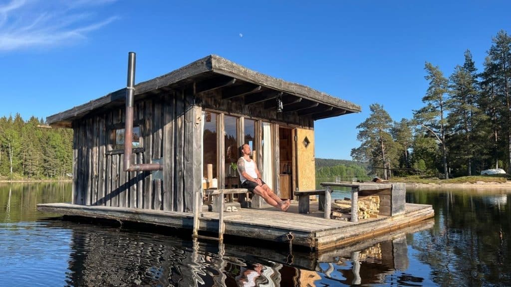 Flytende hytte på en plattform i stille vann, omgitt av skog og blå himmel, med person som nyter utsikten.